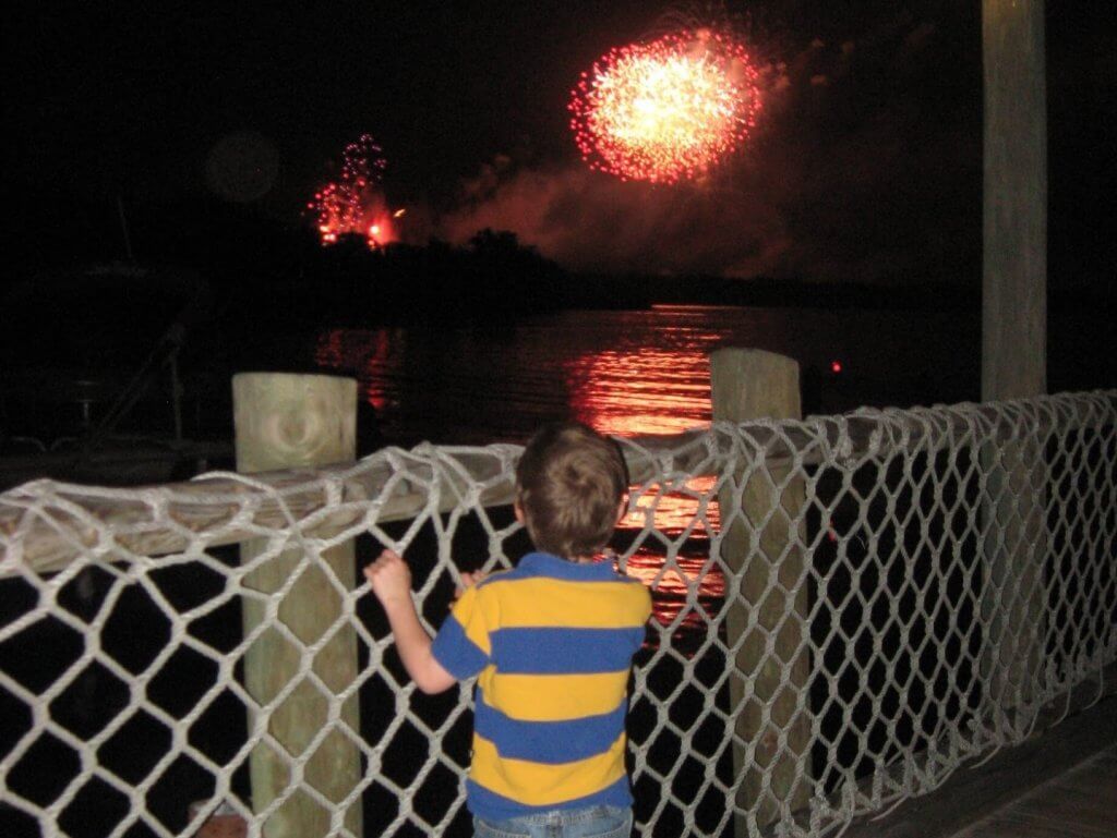 boy at a fence watching fireworks over water