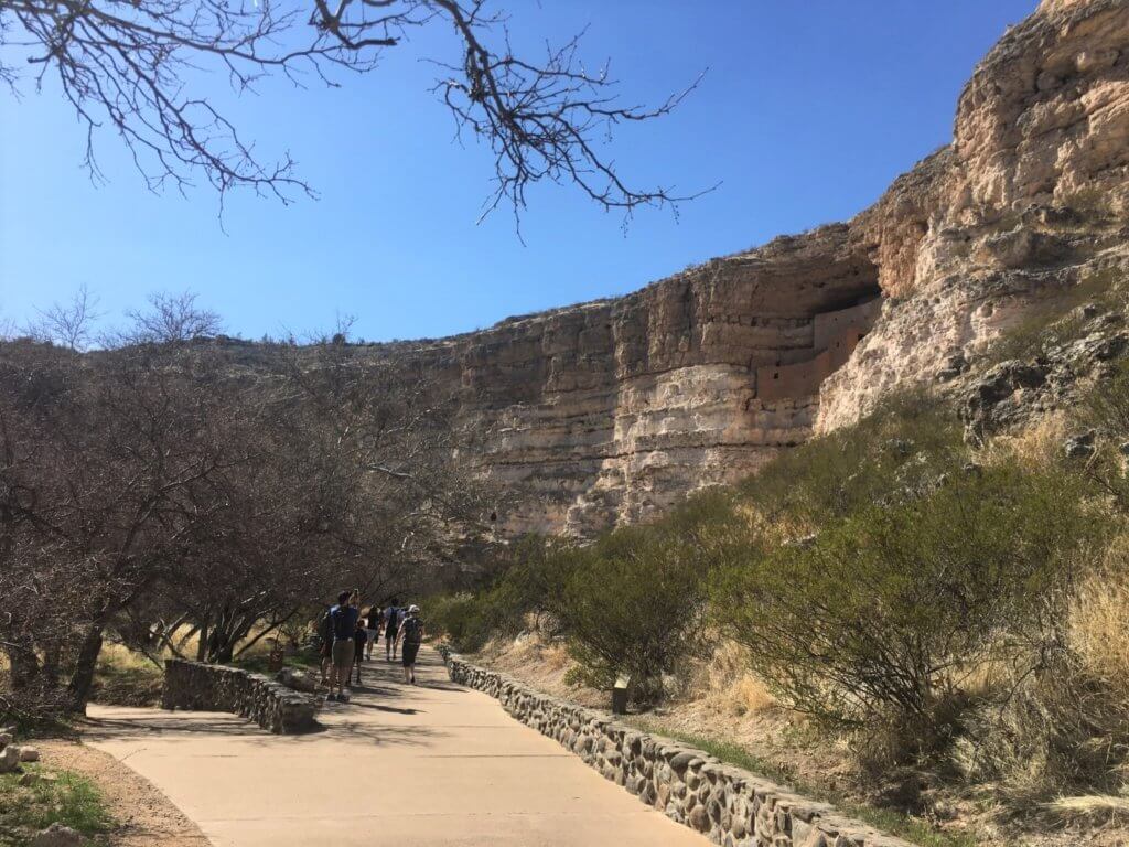 people on sidewalk with rock structure in cliff face